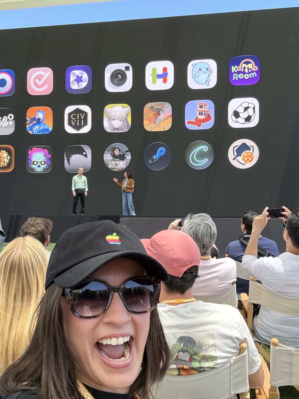 Lilly in an Apple cap in the audience at Apple Park, with the Hearing Buddy whale icon among the app icons on the screen behind her